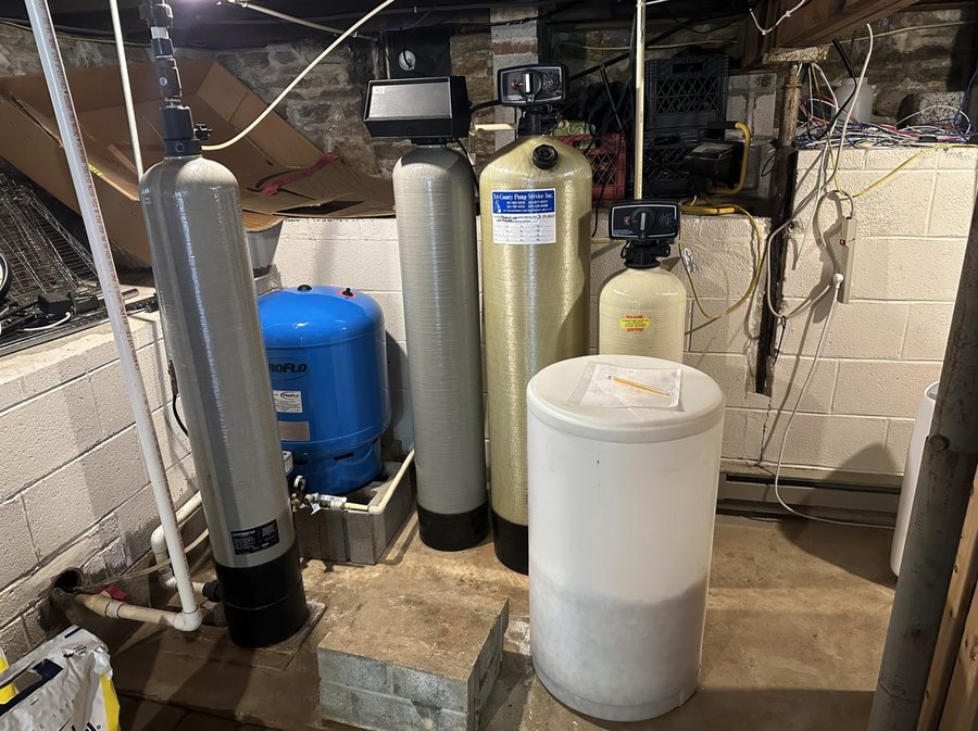 A stacked lineup of sediment filter, well tank, softener, and iron filter tanks in a Falling Waters basement, added one at a time by different companies over the years